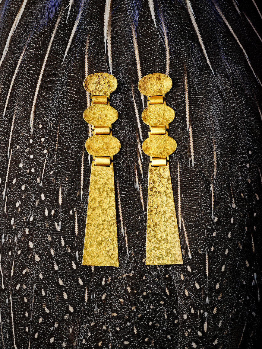 A pair of textured gold drop earrings with stacked oval and rectangular elements, displayed against a dark feather background with blue and black tones.